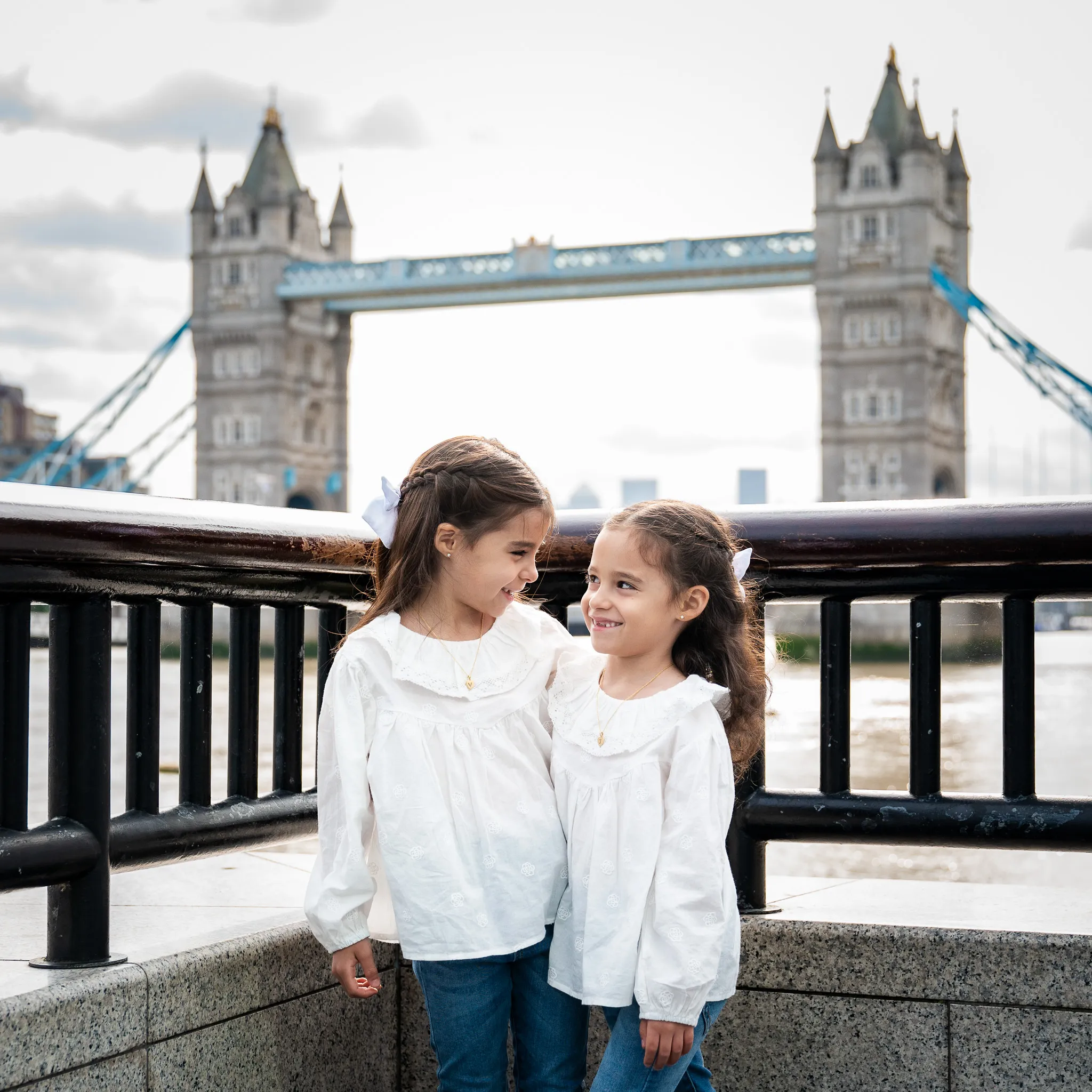 Two little girls in white shirts stand by a railing, with Tower Bridge in the background under a cloudy sky. Central London family photoshoot