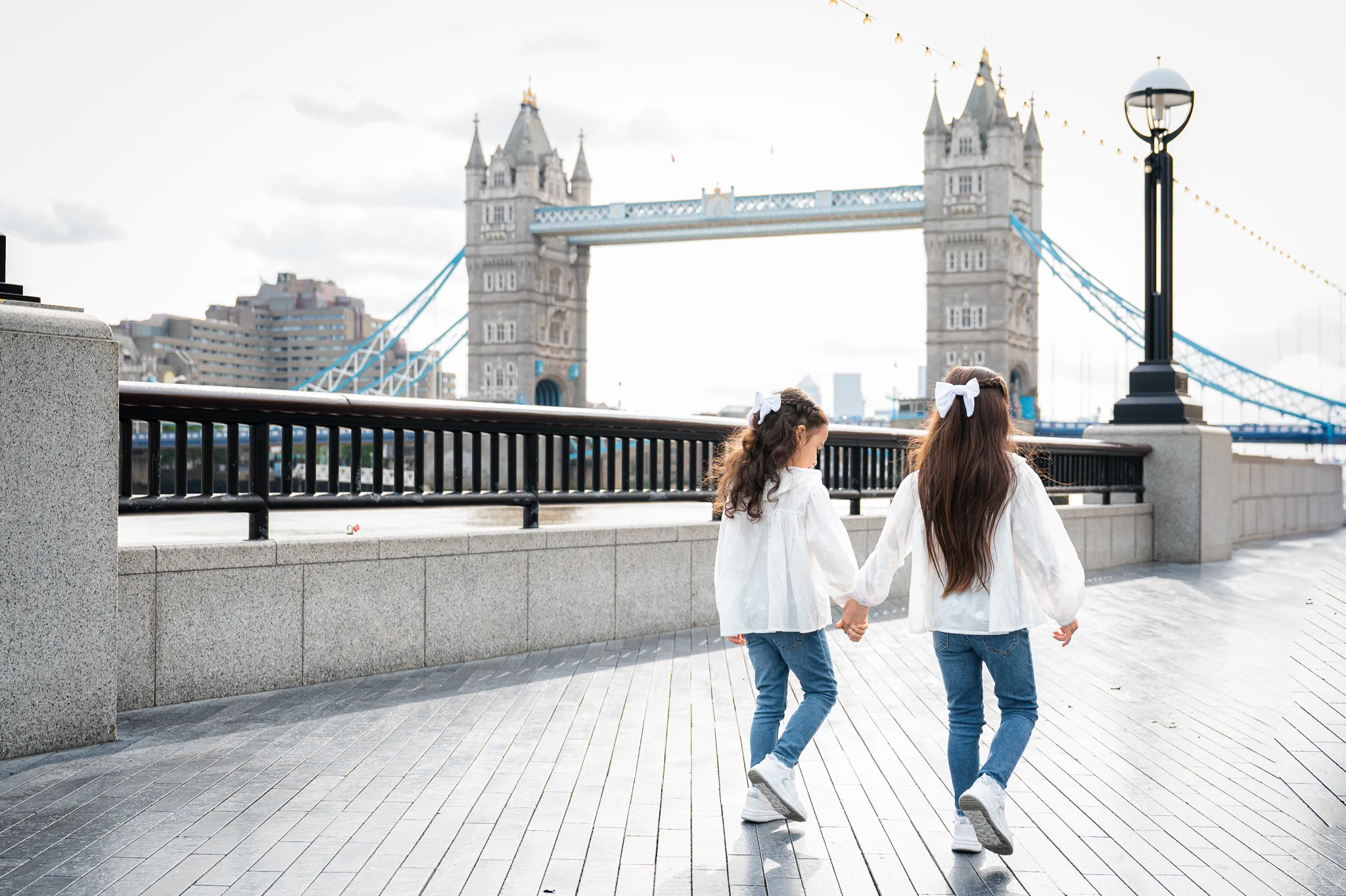 Two girls in white shirts and jeans walk hand-in-hand along the Thames, with Tower Bridge in the background.