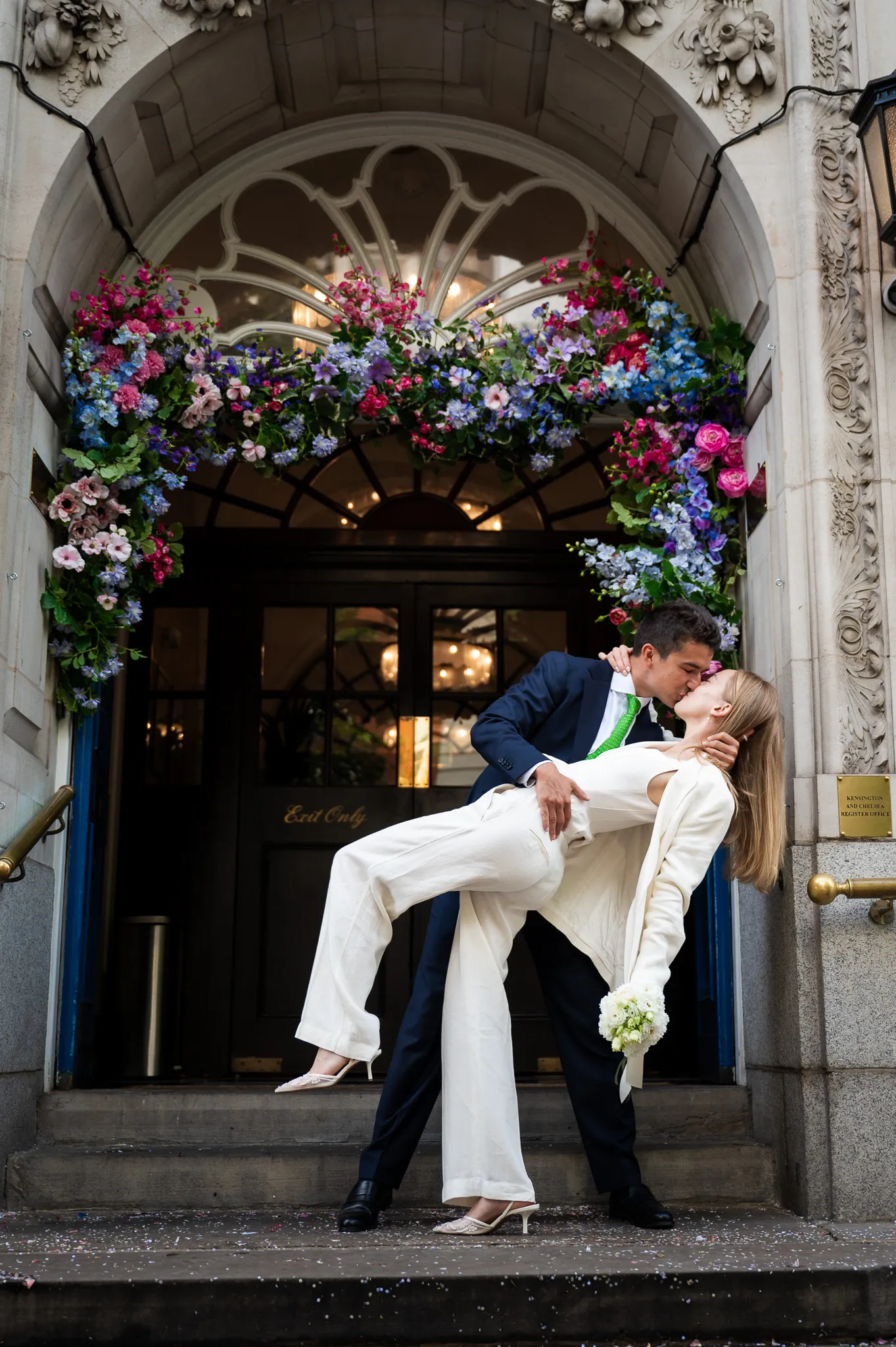 A joyful couple shares a kiss in front of an ornate building entrance adorned with colorful flowers