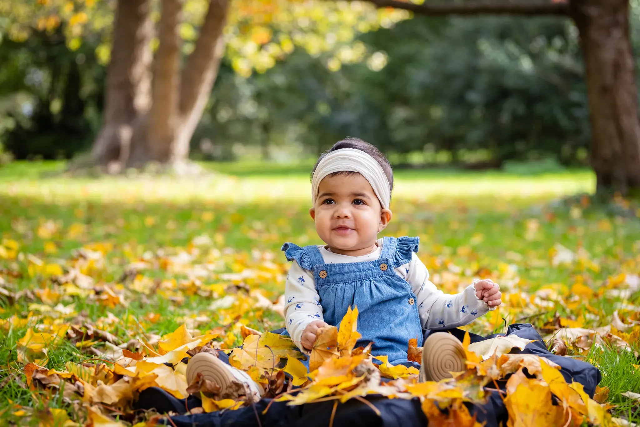 Toddler covered in autumn leaves, sitting on the ground, posing for a quick photoshoot for a family in a park in London