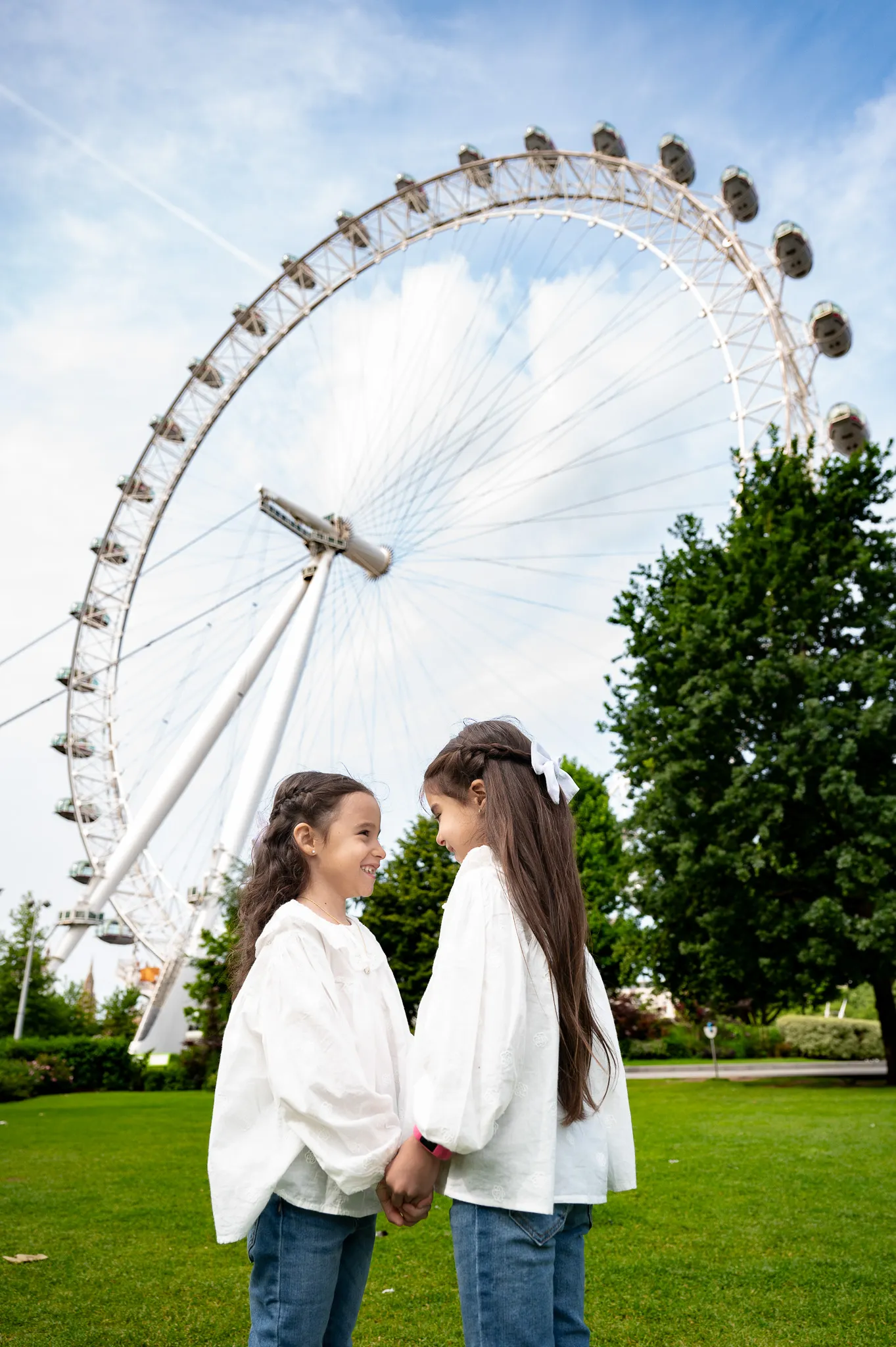 Two girls in white shirts stand hand in hand on green grass, with London eye and blue sky in the background.