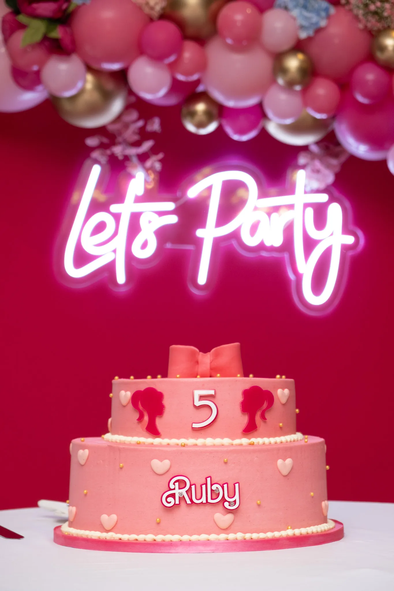 A pink birthday cake in-front of a let's party banner and balloons