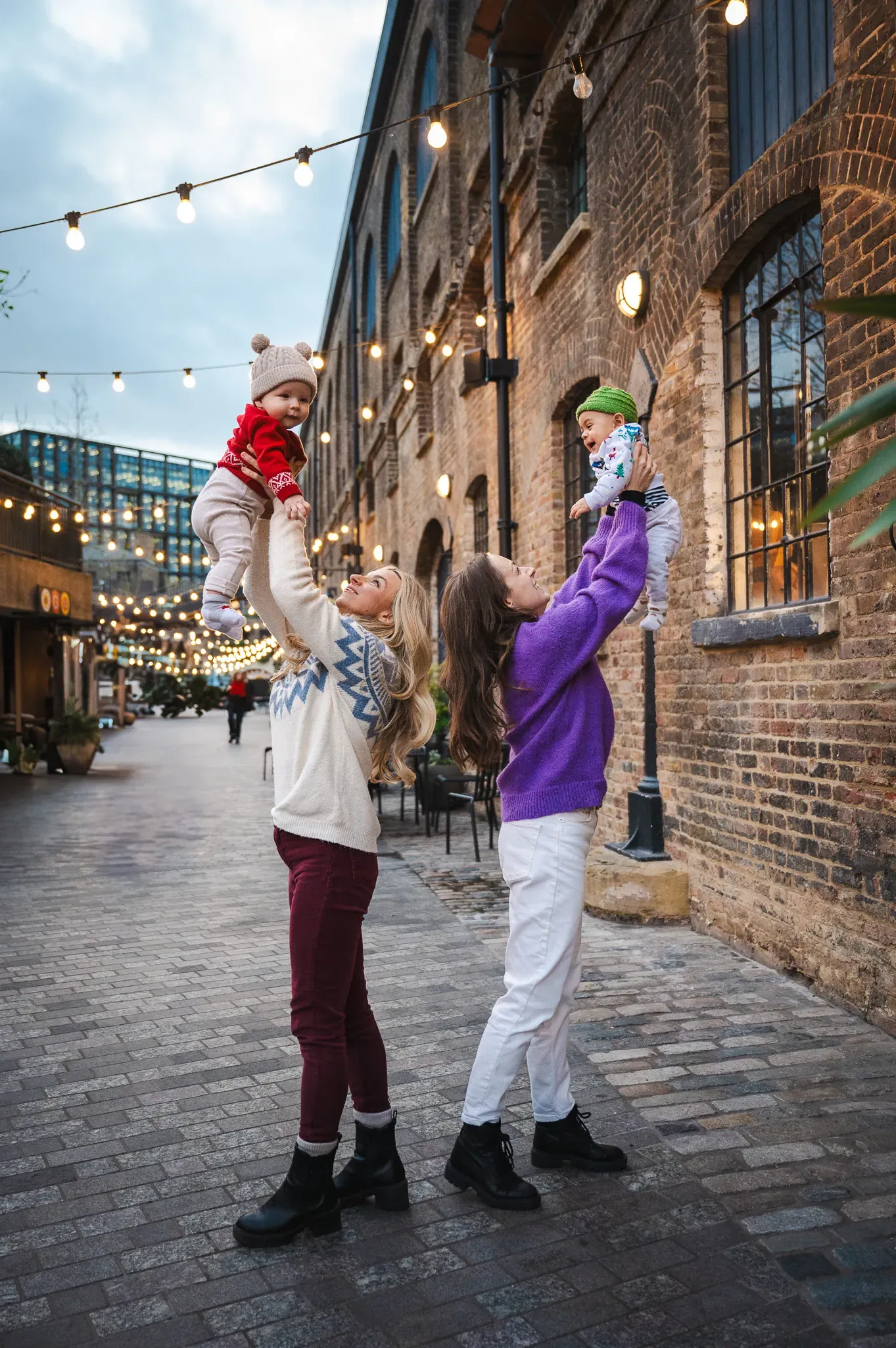 Two mothers with their children, holding their babies and lifting them in the air, posing for a family photoshoot