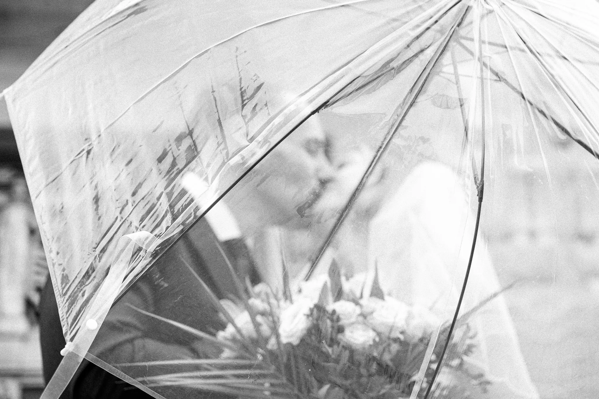 A couple shares a kiss under a clear umbrella, holding a bouquet of flowers
