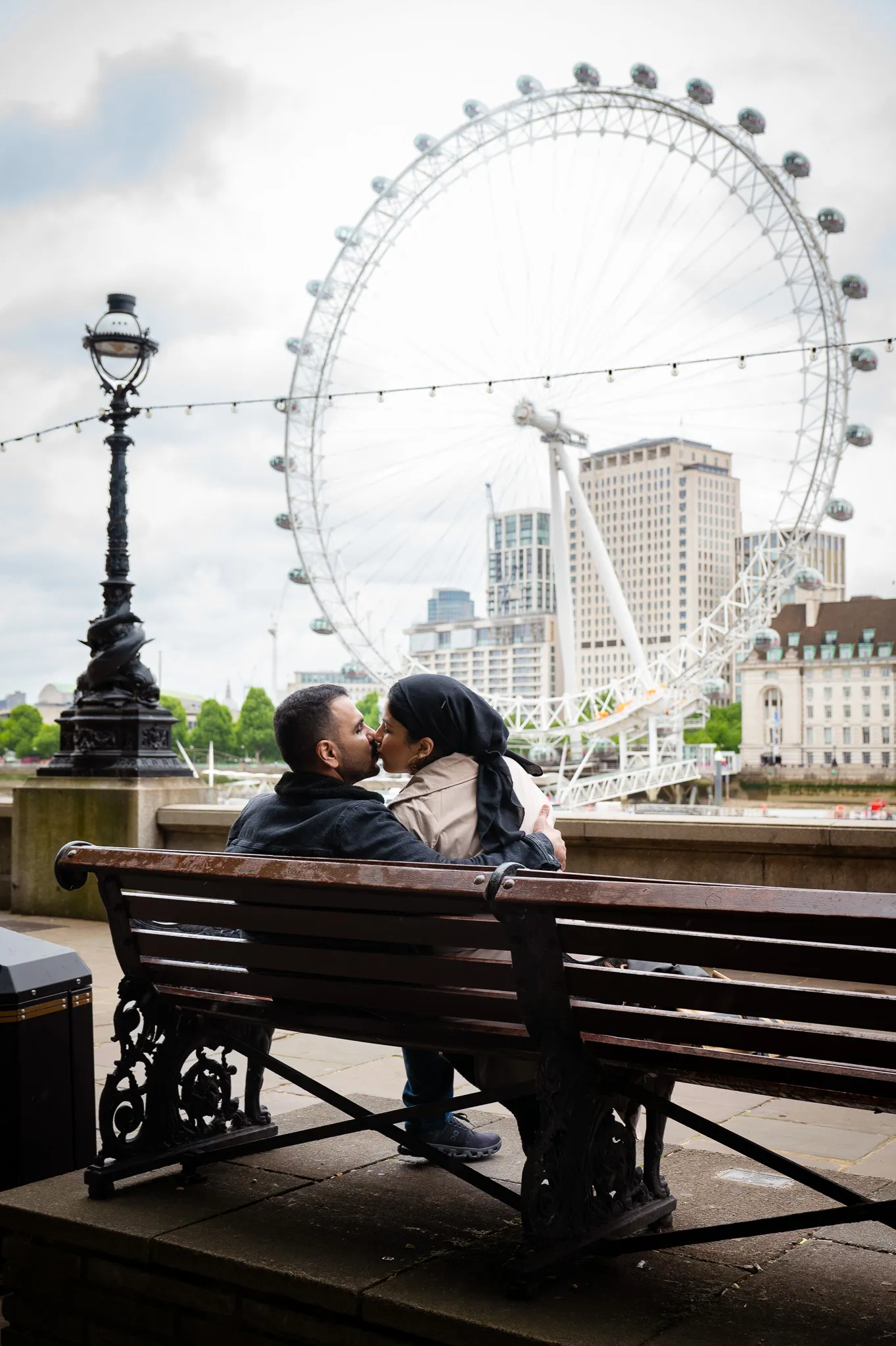A love couple kissing on a bench in front of the London Eye, doing a maternity photoshoot in London
