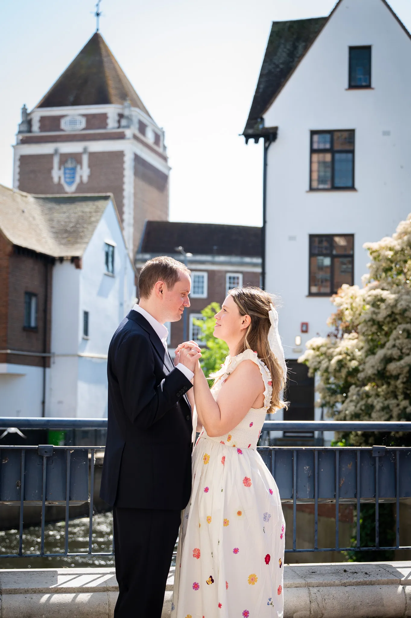 Couple holding hands and gazing at each other on a sunny bridge in front of historic buildings