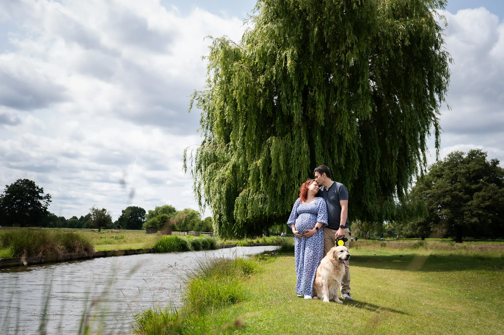 Expecting couple kissing by the river with their golden retriever
