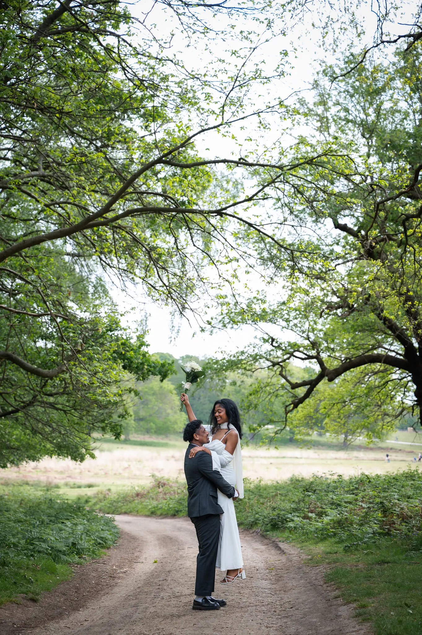 Groom lifting his bride on a country path under tall trees as she raises her bouquet