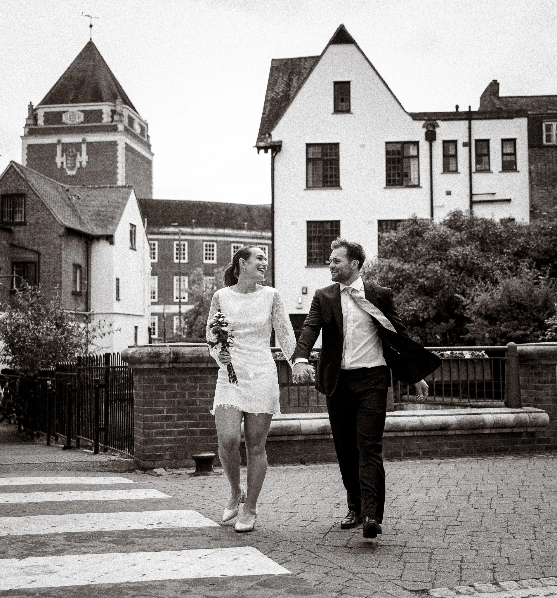 Black and white photo of a newly married couple laughing as they walk hand in hand across a street