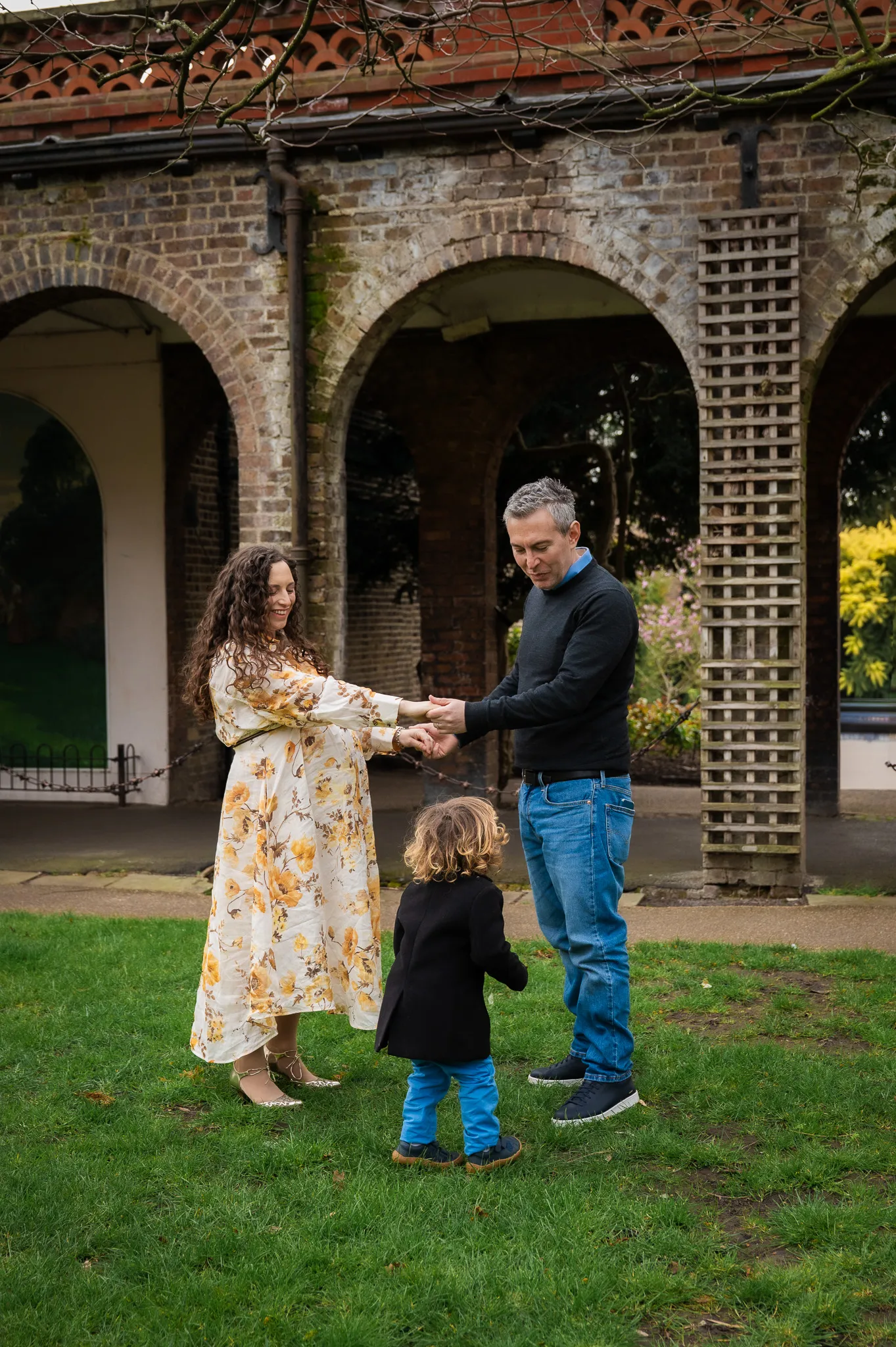 Pregnant mum and dad holding hands with their toddler under brick arches
