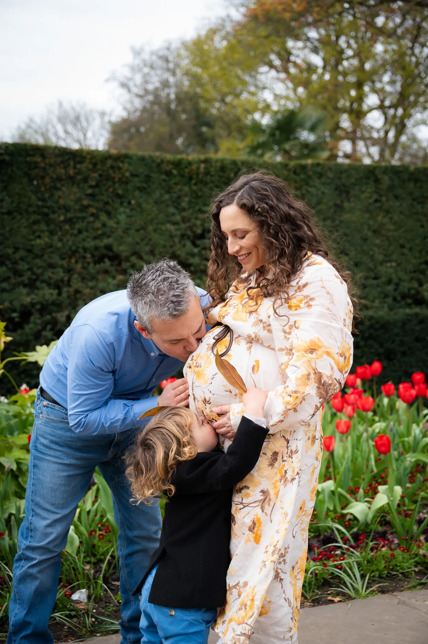 Dad and toddler kissing mum's baby bump beside red tulips