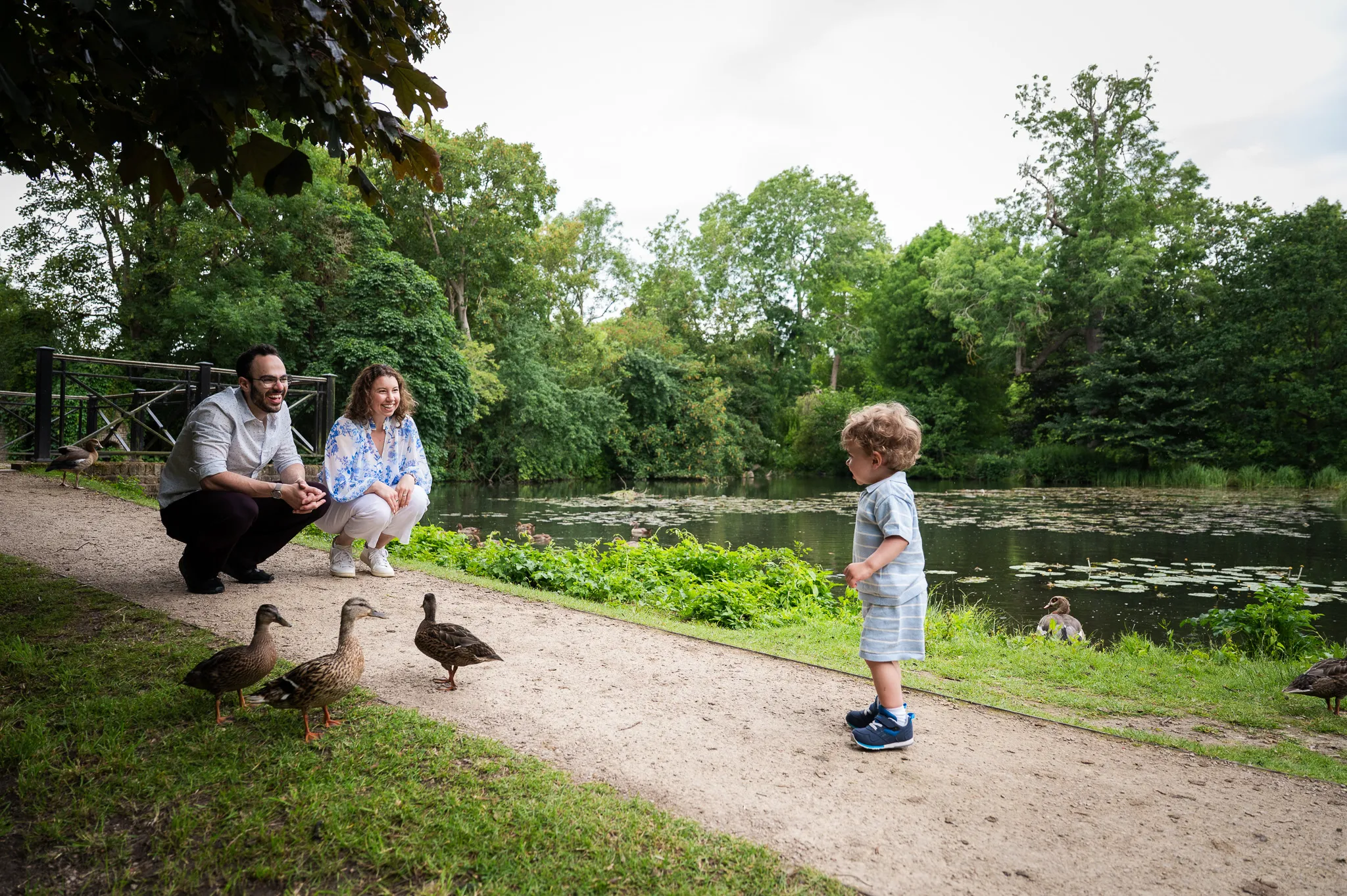 Parents smiling as their toddler meets the ducks by a pond in the park