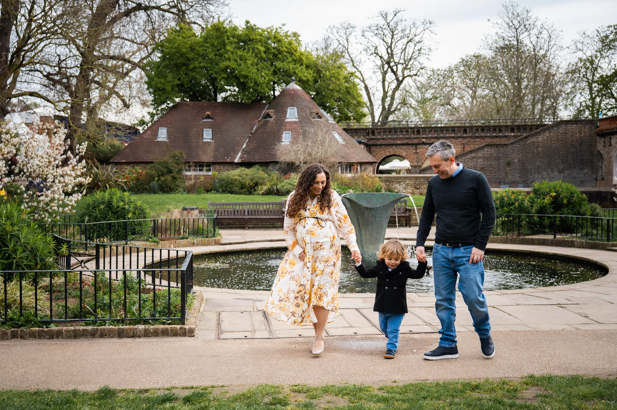Expecting parents walking hand in hand with their toddler by a garden pond