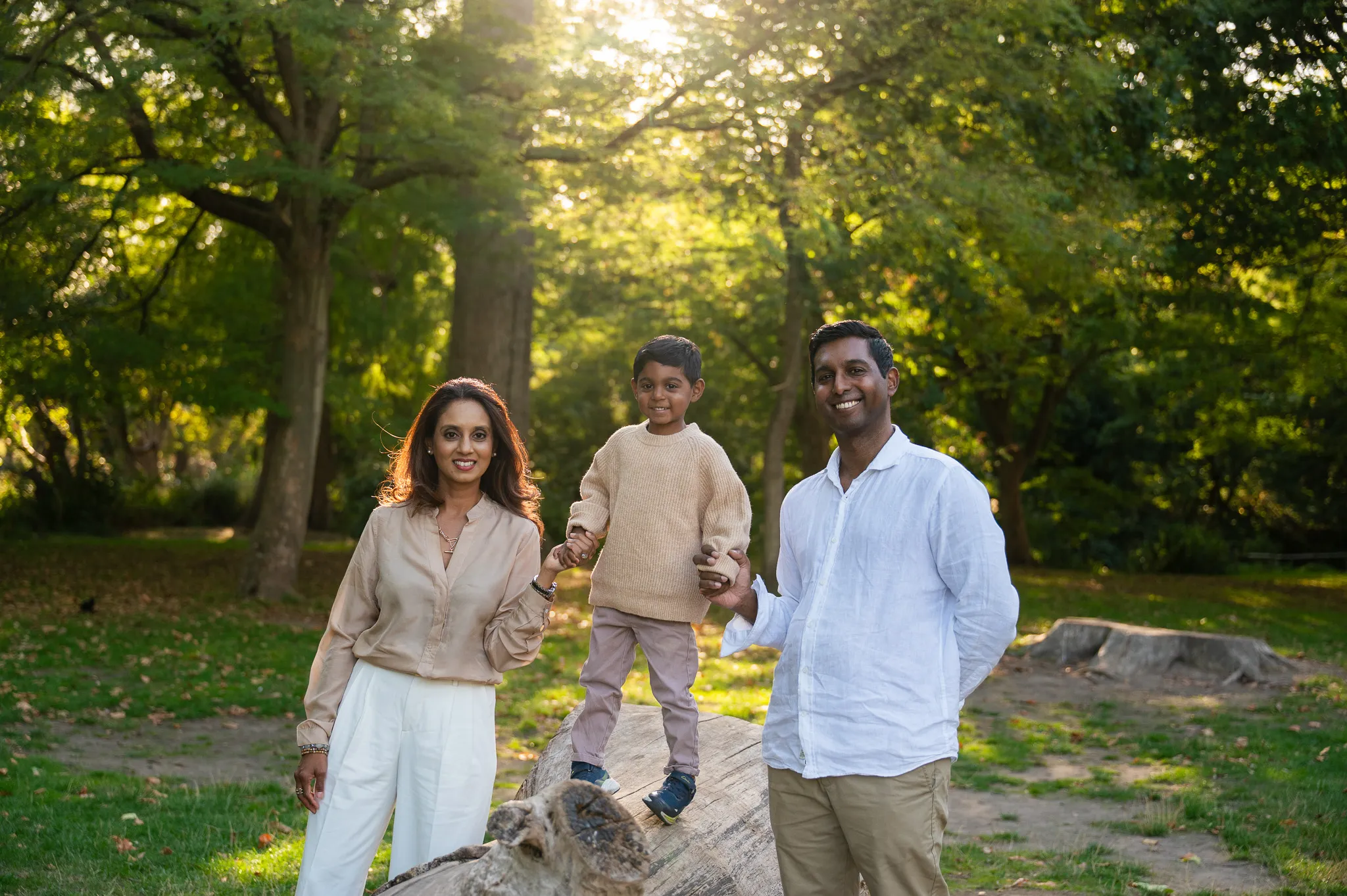 Smiling family of three holding hands with their son as he balances on a fallen log in a sunlit park