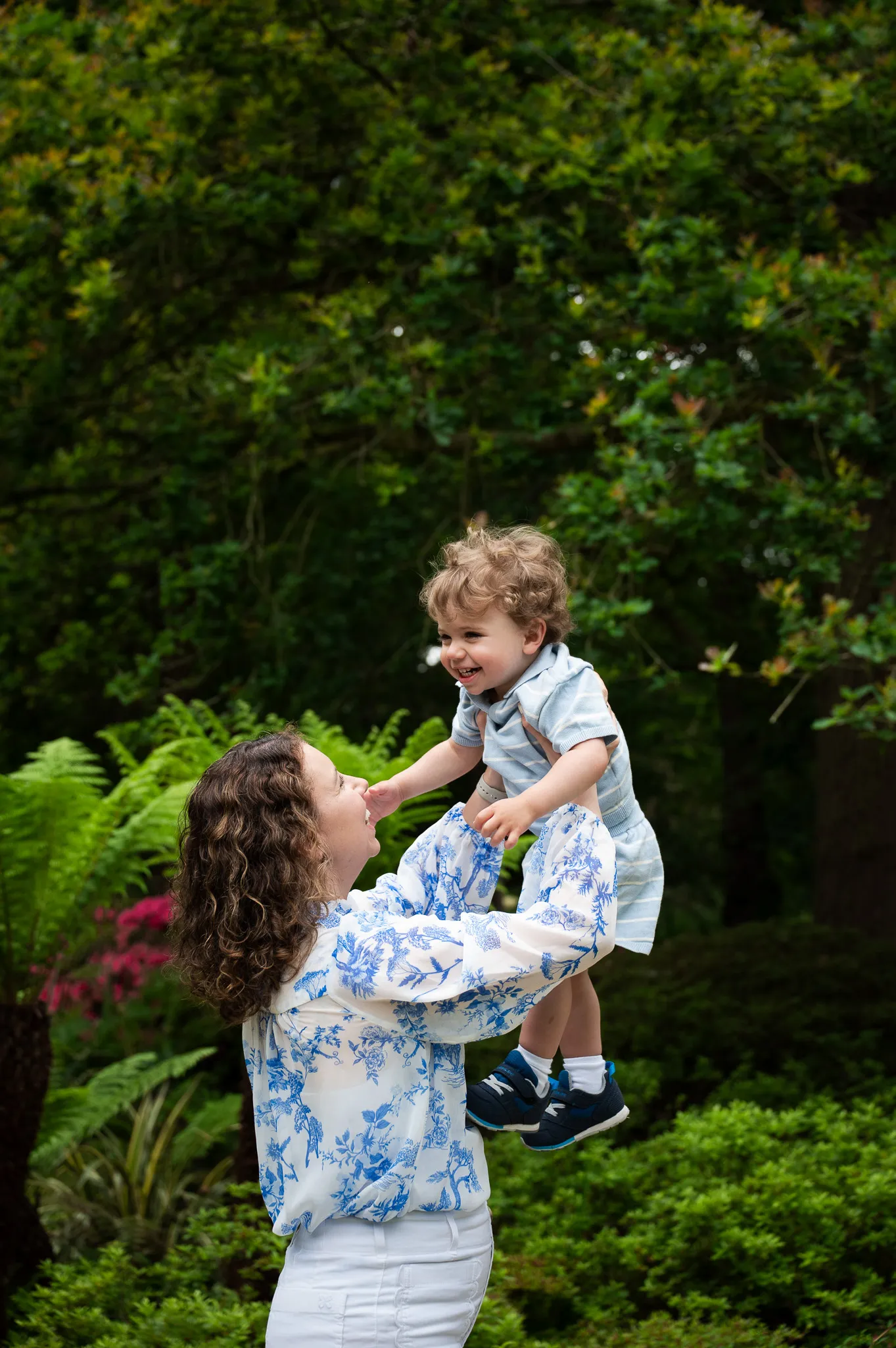 Mum lifting her laughing toddler up high among ferns and flowers