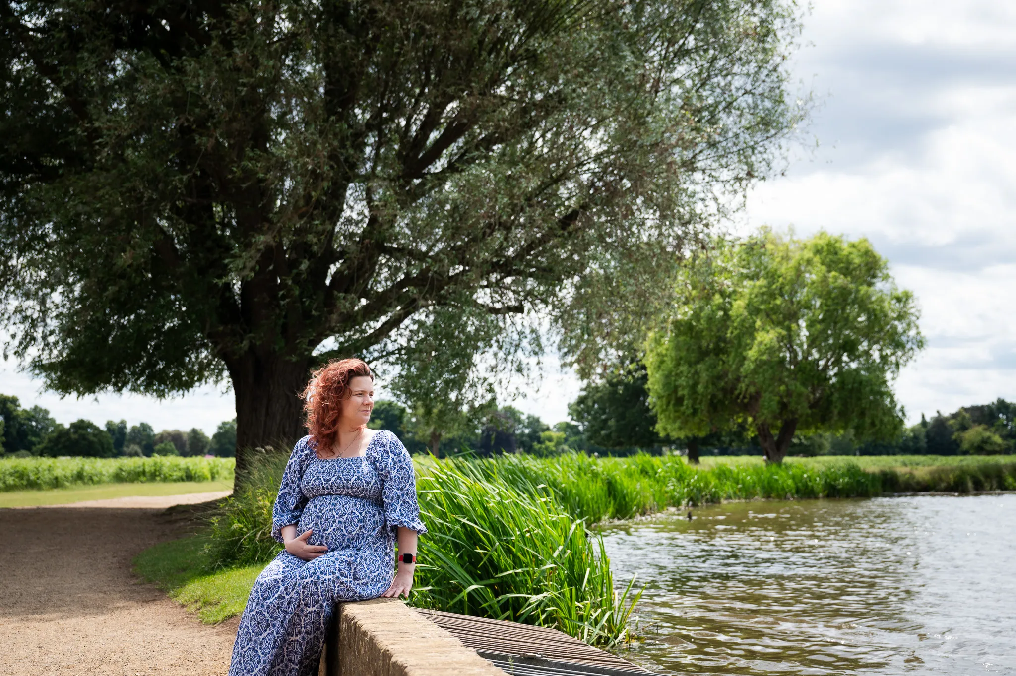 Pregnant mum in a blue floral dress sitting by the river holding her bump