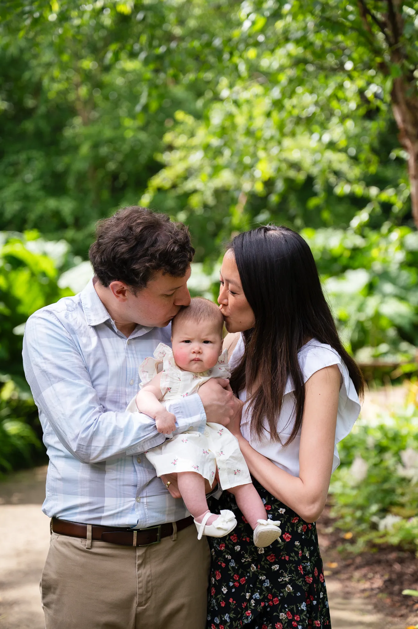 Parents kissing their baby daughter on both cheeks in a garden