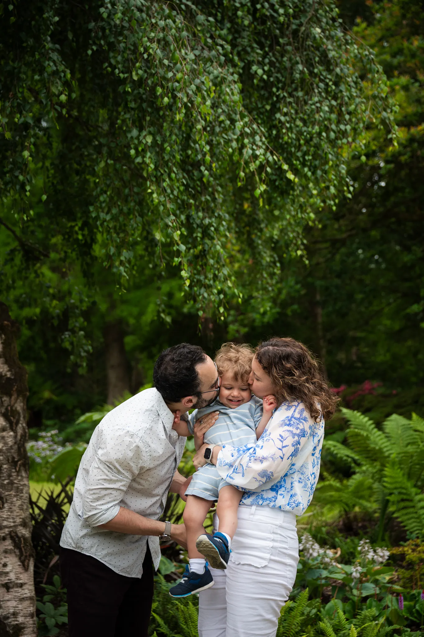 Parents kissing their toddler on both cheeks under a willow tree