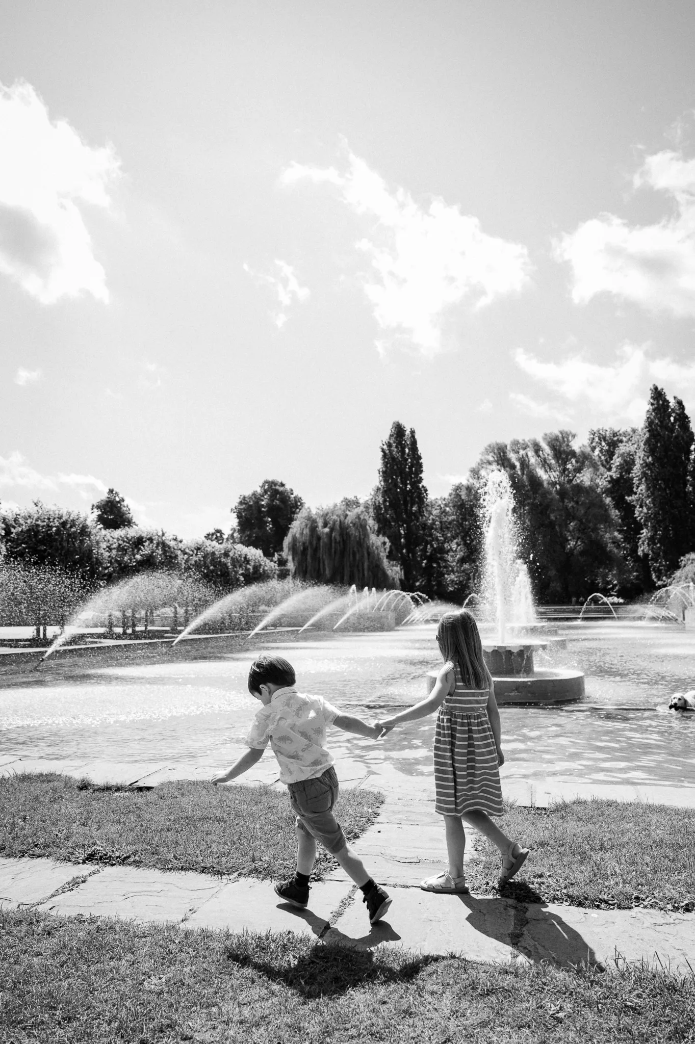 Black and white photo of a brother and sister running hand in hand by the fountains