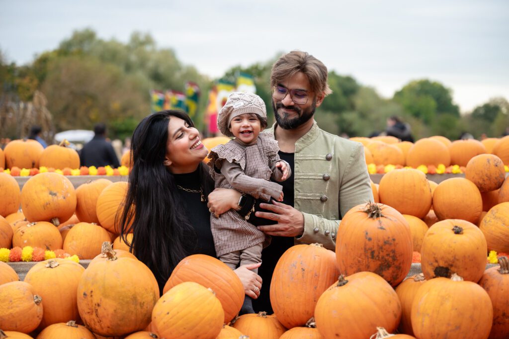 A family of three smiles while sitting among numerous pumpkins, with trees and colourful flags in the background.